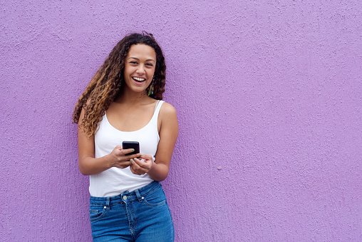 Cheerful young woman with a mobile phone standing against wall with copy space