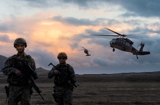 Two soldiers walking in the battlefield while two military helicopter flying over them during a military operation at dusk.