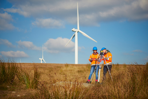 two wind farm engineers using a builder's level to plan out the expansion of the wind farm site. they are wearing orange hi vis jackets and blue hard hats . one is male , one is female. In the background wind turbines can be seen across the landscape.