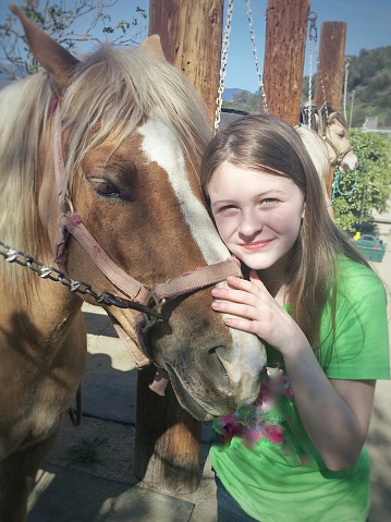 Girl wearing green shirt hugging horse head. Cream and tan colored horse harnessed and chain hitched to rustic western posts, Mountains and other horse in background.