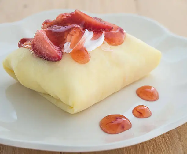 Strawberry crepe cake in white dish placed on wooden floor.Focus strawberry top cake.