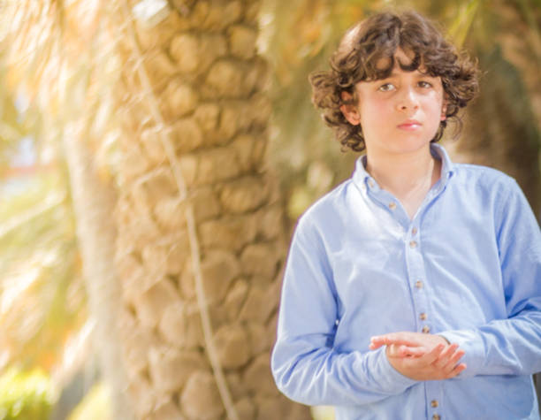 Boy wearing cap and light-colored shirt observes tropical palm tree in park with curiosity. The scene emphasizes his fascination with nature and greenery on bright day.