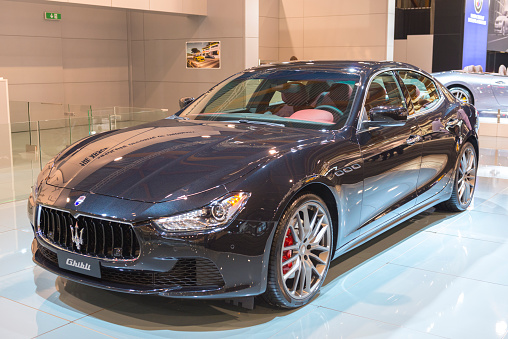 Brussels, Belgium - Januari 12, 2016: Maserati Ghibli luxury saloon car front view. The Maserati Ghibli shares the architecture of the sixth-generation Maserati Quattroporte and is fitted with a 2,979 cc V6 Twin Turbo engine. The car is on display during the 2016 Brussels Motor Show. The car is displayed on a motor show stand, with lights reflecting off of the body. There are people looking around in the background.