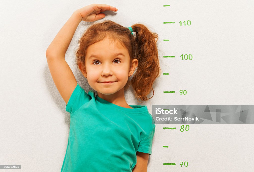 Close portrait of a girl show height on wall scale Close portrait of little girl standing by the scale drawn on the wall in living room and measure her height with hand Child Stock Photo