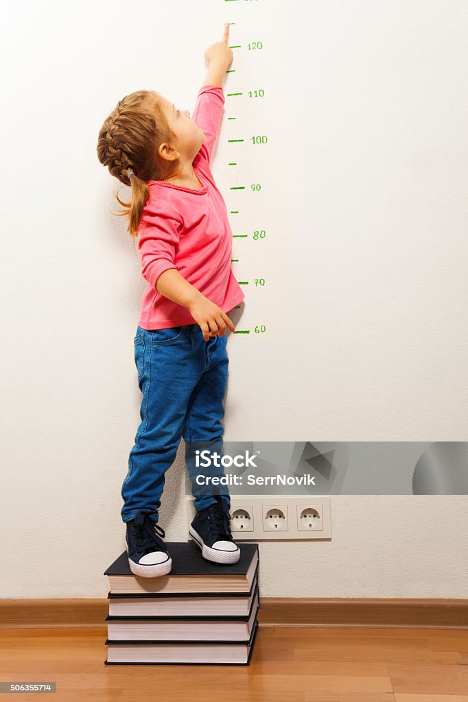 Girl checking height on growth chart at four books Funny girl measuring her height supporting at four big books pointing to the ruler with her finger Child Stock Photo