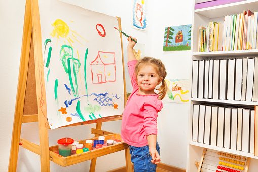 The pretty little girl's cheerful painting on the white canvas at the easel