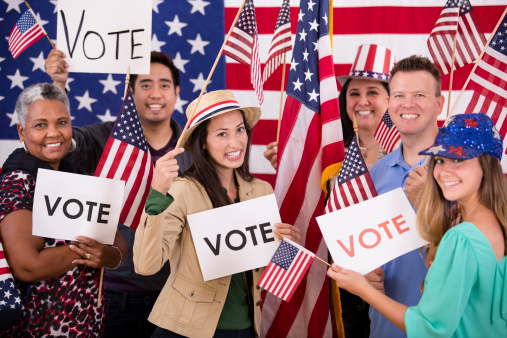 Multi-ethnic and mixed ages group excitedly encourage the public to vote during a political rally. American flags, voting signs.  Primary and presidential elections. 