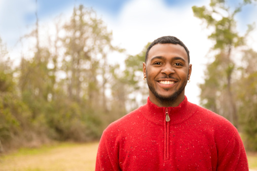 Young adult, African descent man outside at a local park wearing red sweater in autumn season.  Happy, smiling.  Beard.