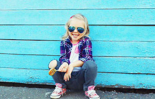 Stylish child with sweet lollipop in city over colorful blue background