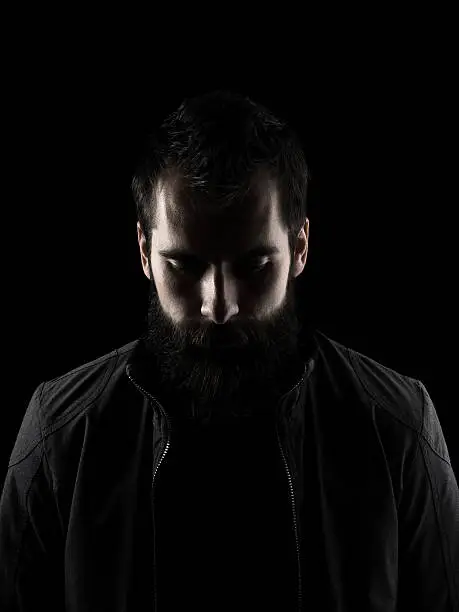 Sad bearded man looking down. High contrast low key dark shadow portrait isolated over black background.