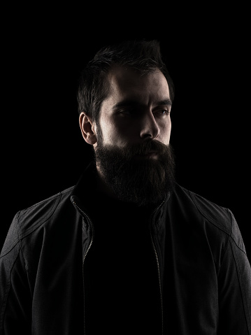 Desaturated serious bearded hipster looking away. High contrast low key dark shadow portrait isolated over black background.