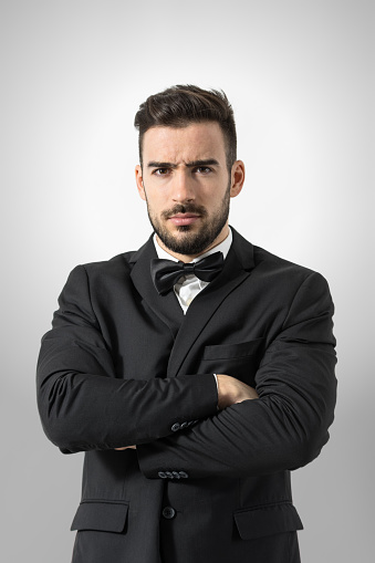 Angry bossy man in tuxedo with crossed arms intense looking at camera. Portrait over gray studio background.