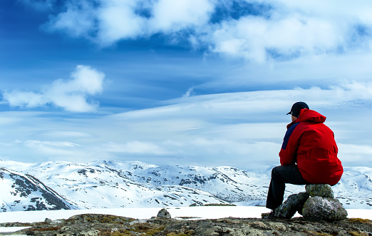 Man sitting on rocks and looking at the snow-covered hills. Blue sky with clouds. Norway