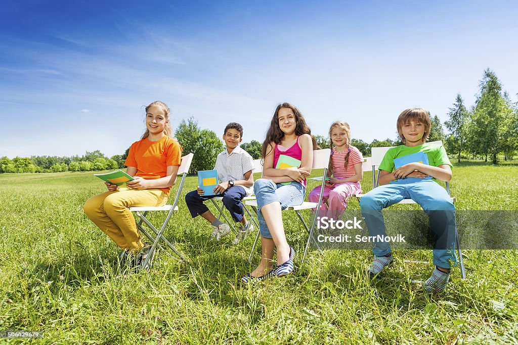 School children hold notebooks and sit on chairs Smiling school children hold exercise books and sit on chairs like in school outside in summer time African Ethnicity Stock Photo