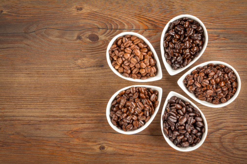 Overhead view of a variety of fresh roasted coffee beans in petal shaped bowls on a brown wooden background with copy space.