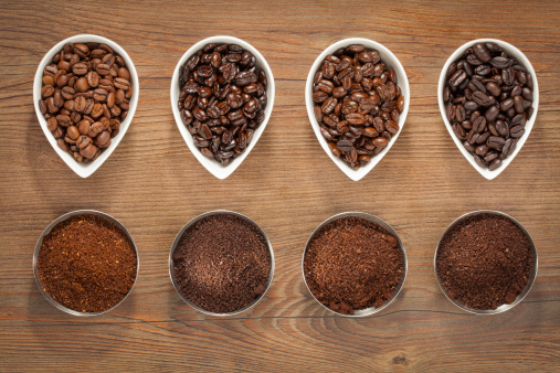 Overhead view of four varieties of roasted coffee beans with their freshly ground counterparts in small bowls on a brown wooden background.