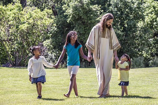 Jesus Christ is walking in an open grass meadow holding hands with a teenager and her two younger sisters. All three ethnically diverse girls are listening and looking up at Jesus as he talks and teaches and leads them forward.