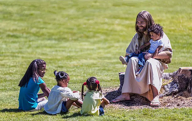 Jesus Christ is sitting in a sunny summer open meadow holding a squirming baby boy as he talks to a teenager and two younger girls sitting on the grass in front of him. The four children are ethnically diverse siblings with African, Latin American and Hispanic roots. They are trying to pay attention to Jesus as he talks and teaches them.