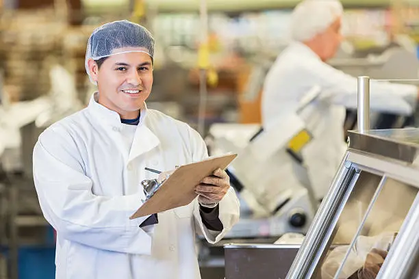 Mid adult Hispanic man is looking at the camera and smiling. He is using a clipboard to take orders from customers in local grocery store or supermarket. Butcher or deli manager is standing behind deli counter. He is wearing a chef's coat and hair net.