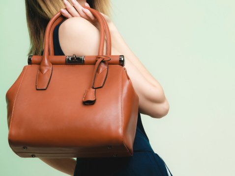 Elegant outfit. Closeup of brown leather bag handbag in hand of stylish woman fashionable girl on green. Female fashion.