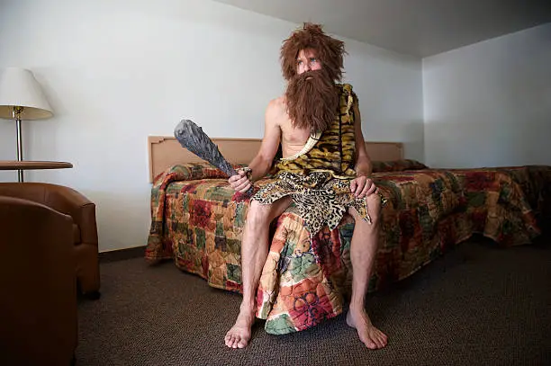 Traveling caveman sitting with a club on bed in old-fashioned motel room