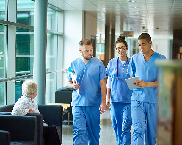 junior doctors three junior doctors walking along a hospital corridor discussing case and wearing scrubs. A patient or visitor is sitting in the corridor as they walk past . medical student photos stock pictures, royalty-free photos & images