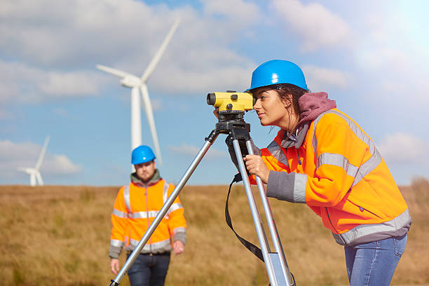 female windfarm engineer two wind farm engineers using a builder's level to plan out the expansion of the wind farm site. they are wearing orange hi vis jackets and blue hard hats . one is male , one is female. In the foreground the female is looking through the level whilst the male engineer is approaching .In the background wind turbines can be seen across the landscape. civil engineering photos stock pictures, royalty-free photos & images