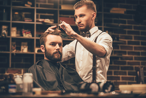 Young bearded man getting haircut by hairdresser while sitting in chair at barbershop