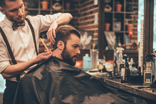 Young bearded man getting haircut with straight edge razor by hairdresser while sitting in chair at barbershop