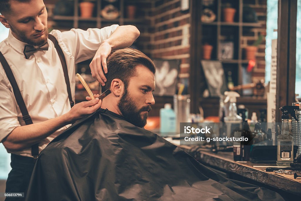 Making hair look magical. Young bearded man getting haircut with straight edge razor by hairdresser while sitting in chair at barbershop Barber Shop Stock Photo