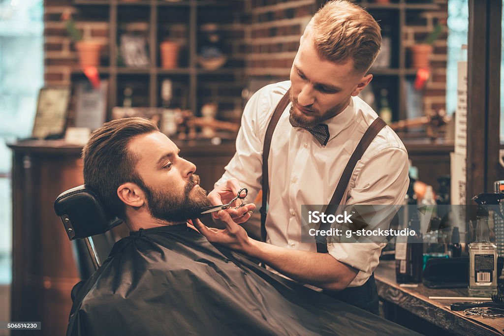 Beard grooming. Side view of young bearded man getting beard haircut by hairdresser while sitting in chair at barbershop Barber Shop Stock Photo