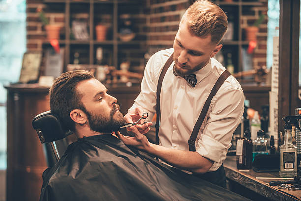 Beard grooming. Side view of young bearded man getting beard haircut by hairdresser while sitting in chair at barbershop barber shop stock pictures, royalty-free photos & images