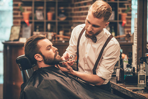 Beard grooming. Side view of young bearded man getting beard haircut by hairdresser while sitting in chair at barbershop barber stock pictures, royalty-free photos & images