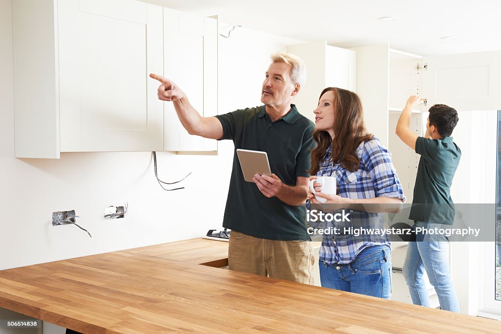 Woman With Carpenter Looking At Plans For Kitchen Woman With Carpenter Looking At Plans For Kitchen On Digital Tablet Quality Control Stock Photo