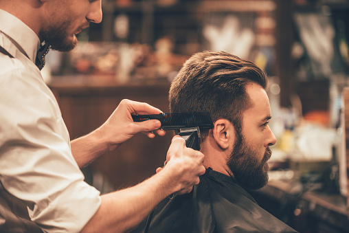 Close up side view of young bearded man getting haircut by hairdresser with electric razor at barbershop