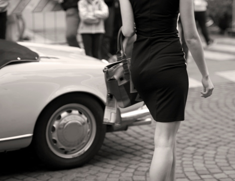 Fashion model and oldtimer on a street of Gorizia, Italy. Unrecognized person wearing small black short dress. On a background is a part of an oldtimer car. Gorizia, Italy, Europe. Black and white image. Selective focus.