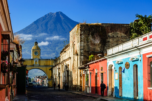Antigua, Guatemala - March 11, 2012: Locals walk to work along Antigua's famous cobblestoned street lined with painted shops, restaurants & hotels. Agua volcano looms behind Santa Catalina Arch, a landmark in this Spanish colonial town & UNESCO World Heritage Site. Originally the arch allowed nuns to pass from one side of Santa Catalina convent to the other without going outside. Ruins of the convent remain to the right of the arch.