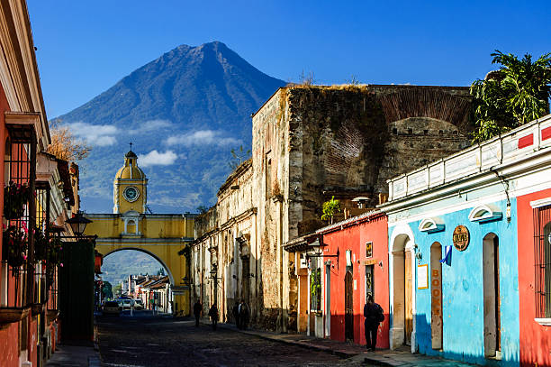 Famous arch and volcano view, Antigua, Guatemala Antigua, Guatemala - March 11, 2012: Locals walk to work along Antigua's famous cobblestoned street lined with painted shops, restaurants & hotels. Agua volcano looms behind Santa Catalina Arch, a landmark in this Spanish colonial town & UNESCO World Heritage Site. Originally the arch allowed nuns to pass from one side of Santa Catalina convent to the other without going outside. Ruins of the convent remain to the right of the arch. agua volcano photos stock pictures, royalty-free photos & images