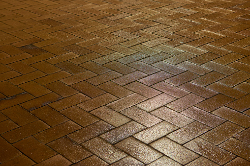 Elevated view of a textured brick sidewalk with a red and yellow color. The sidewalk has a zig-zag pattern and is rough to look at. Shiny wet surface