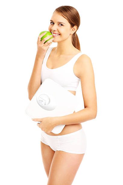 Fit woman with apple and bathroom scales A picture of a fit woman with a green apple and bathroom scales over white background apple women smiling human teeth stock pictures, royalty-free photos & images