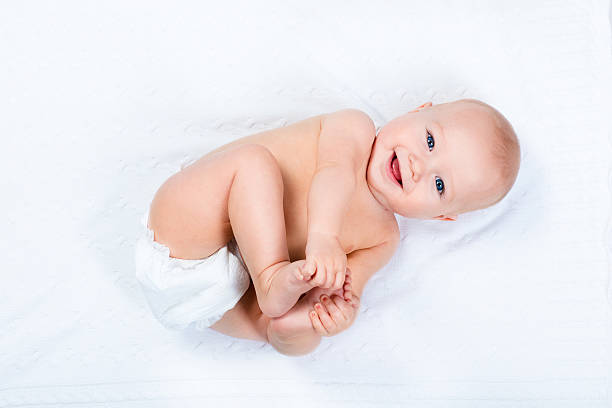 Little baby wearing a diaper Funny little baby wearing a diaper playing on a white knitted blanket in a sunny nursery. Child after bath or shower on a fresh towel. Infant nappy change and skin care. Cute kid playing with his feet taking a bath photos stock pictures, royalty-free photos & images