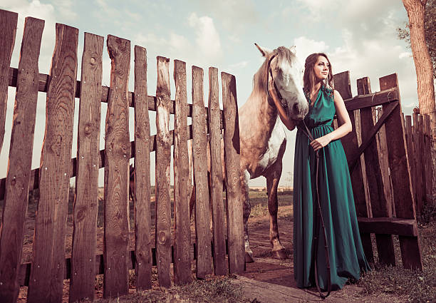 young pretty woman in green dress with white horse at old wooden fence