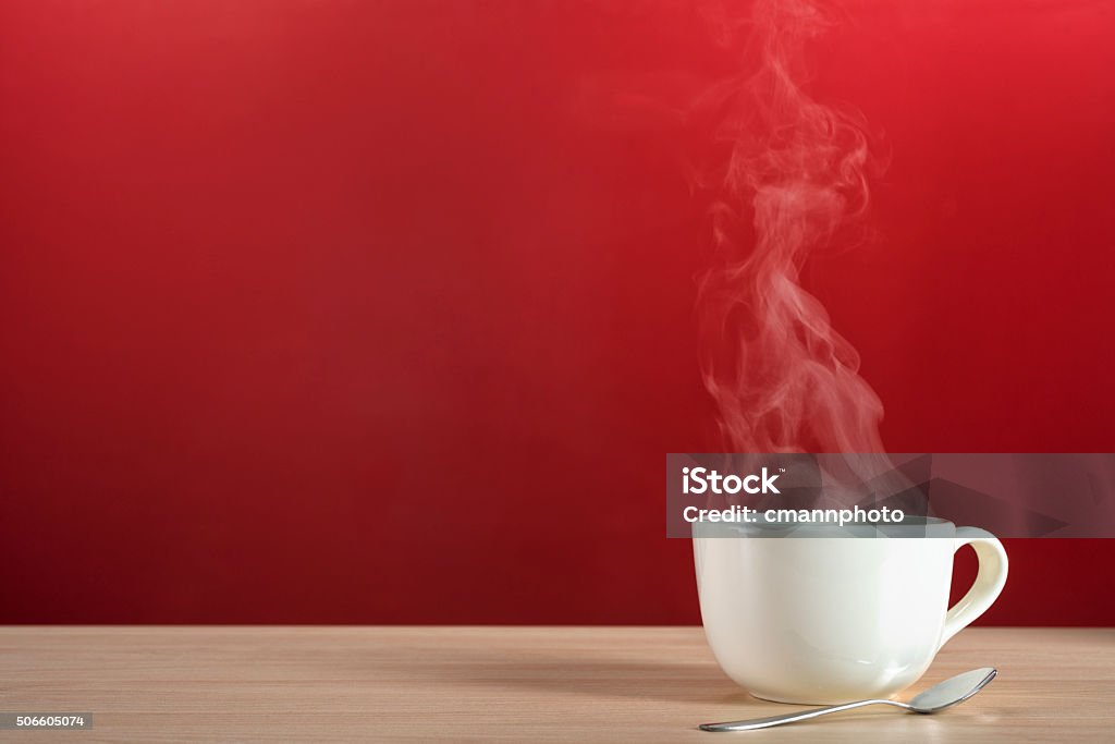 Steamy rising from a large cup of coffee or tea Steam rising from a large white porcelain cup of hot coffee or tea sitting with a silver spoon on a blonde colored wooden table against a red wall Coffee - Drink Stock Photo
