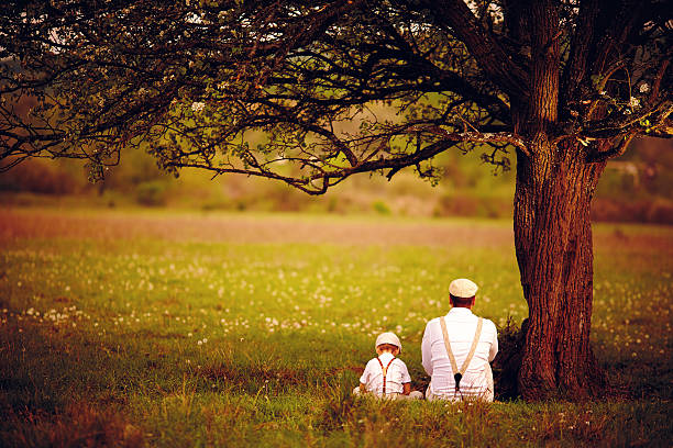 père et fils assis sous l'arbre sur la pelouse de printemps - tree grass family human relationship family photos et images de collection