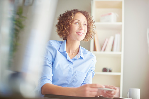 a young woman sits at her desk in a small home office and looks out of the window daydreaming. Strong light floods the shot from the window in front of her desk.