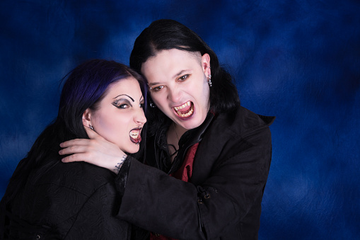 Horizontal colour studio shot on blue of vampire couple closeup. Both in profile with mouths open in snarls, embracing.