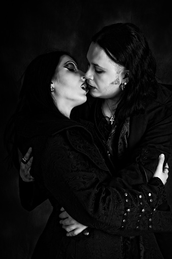 Vertical B&W studio shot with added detail and grain of vampire couple embracing, almost kissing. Waist up black goth clothing. Profile with eyes closed.