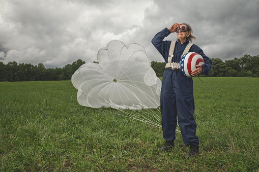Senior lady standing in front of parachute on ground in landing field holding helmet.
