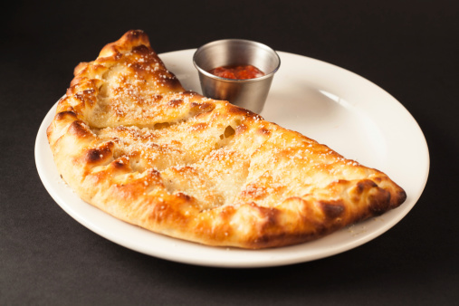 A delicious handmade brick oven Calzone pizza sandwich with marinara sauce composed on a simple black background.