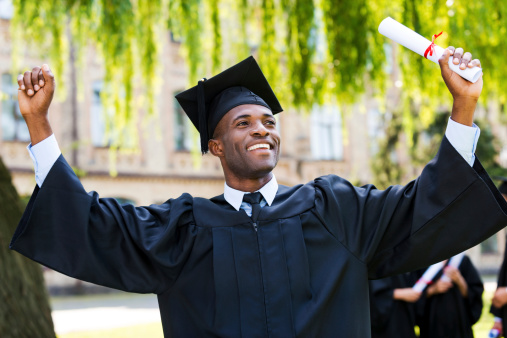 Happy young African man in graduation gowns holding diploma and rising arms up while his friends standing in the background